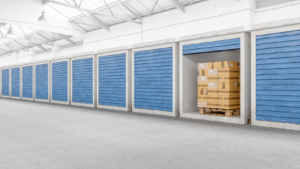 Indoor self-storage facility with multiple blue roll-up doors, one open unit containing stacked cardboard boxes on a pallet.