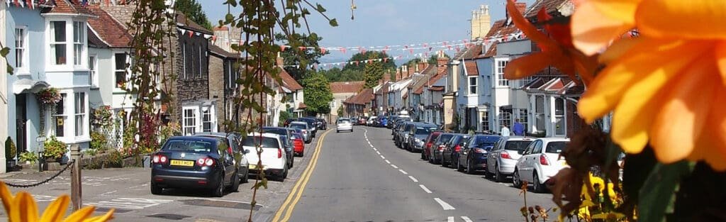 A picturesque English village street lined with parked cars, traditional houses, and festive bunting, framed by bright orange flowers in the foreground.